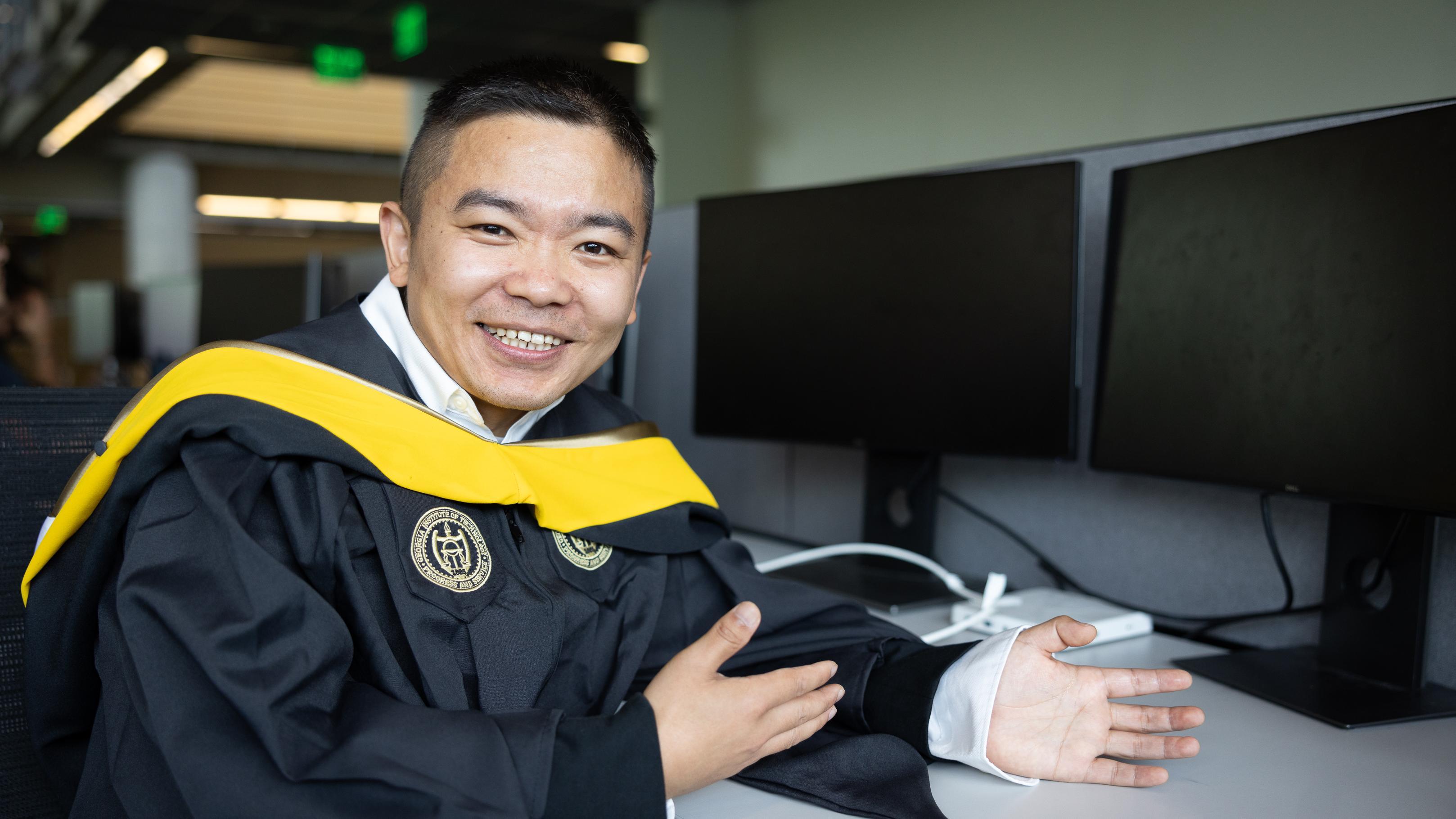 Man in graduation attire sits in front of computers smiling