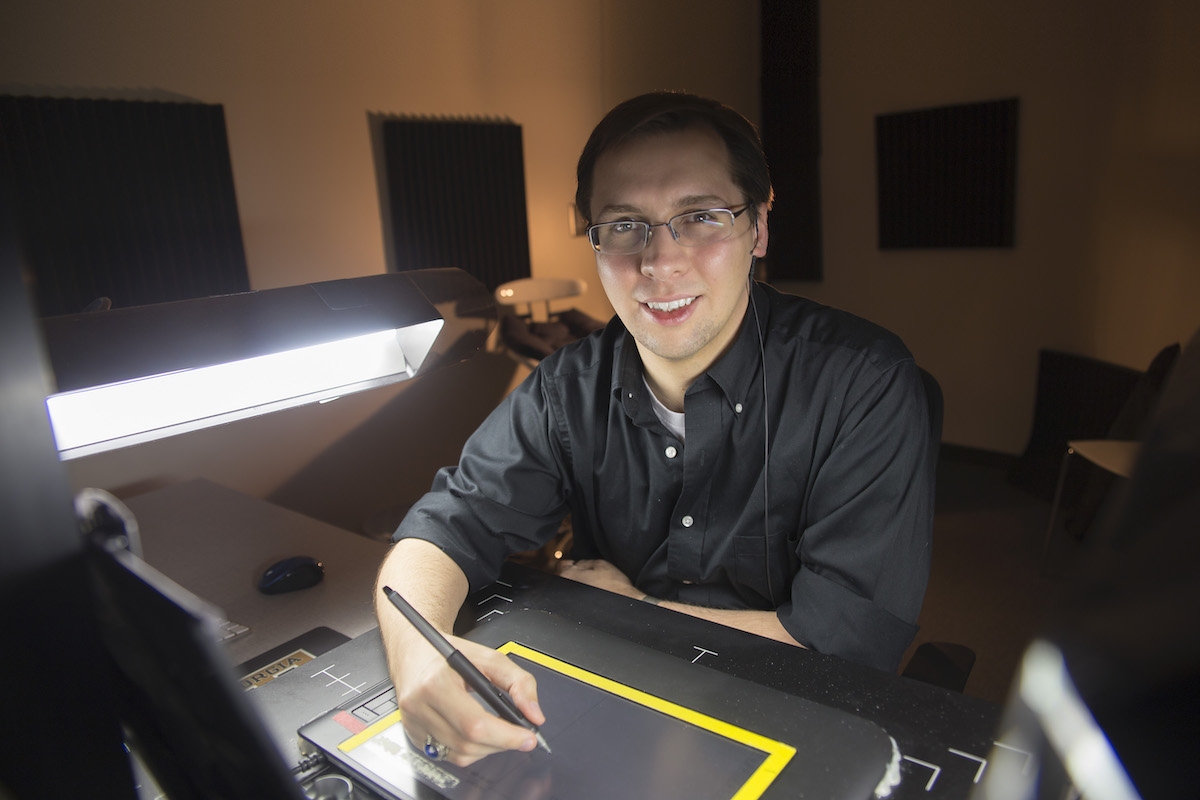 A man wearing glasses sits in front of a computer and smiles