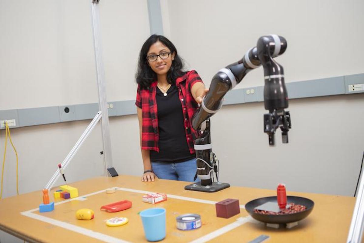 A smiling student stands next to a robotic arm in front of table full of household items