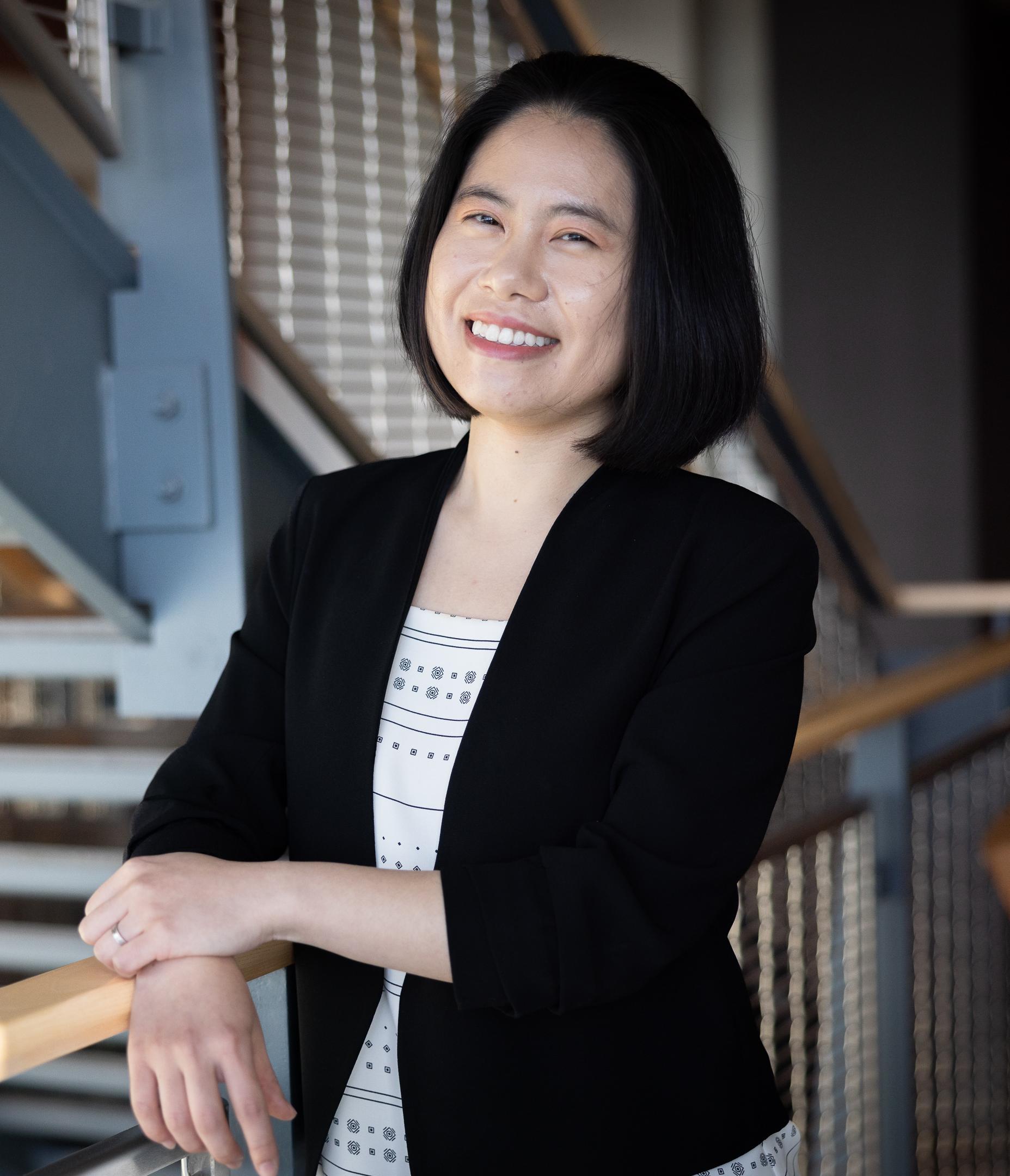A dark haired woman smiles at the camera and poses indoors for a professional photo. She is wearing a dark business jacket with a white shirt underneath. 