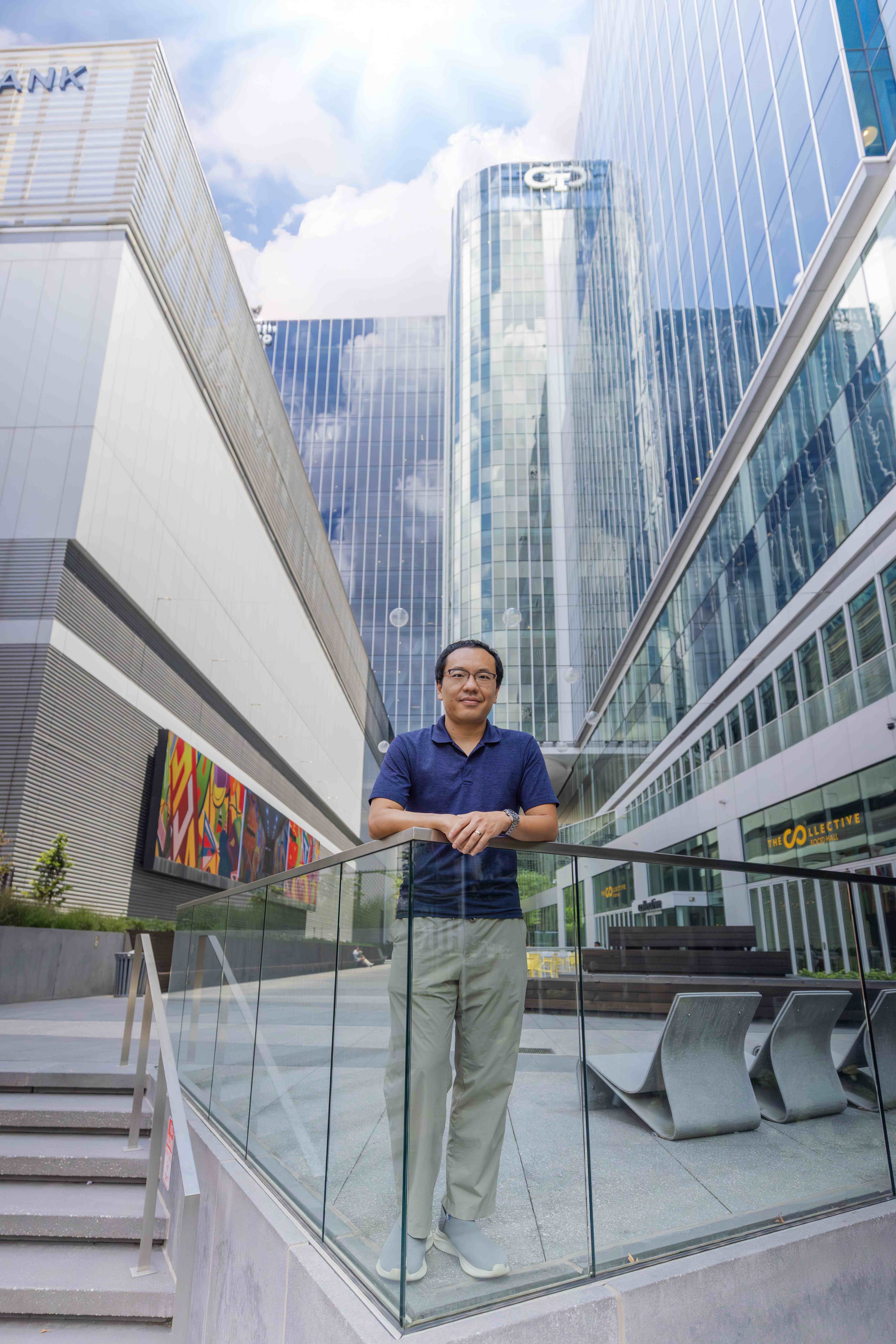 A man standing in the court yard of a tall building. He is leaning on a glass wall and is dressed in a navy blue polo and khaki pants.