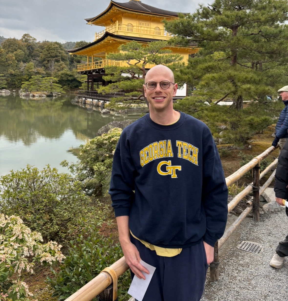 A man standing on a bridge in front of a pond and a traditional styled Japanese house. He is smiling and wearing a Georgia Tech pullover. The color of the house matches his sweatshirt. 