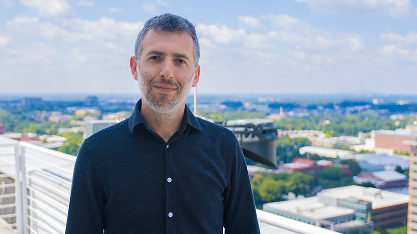A short haired man stands outside on a rooftop balcony. He is wearing a dark blue button up shirt, has a short beard, and is smiling at the camera. There is a blue sky with clouds behind him, and parts of Georgia Tech's campus are out of focus behind him.