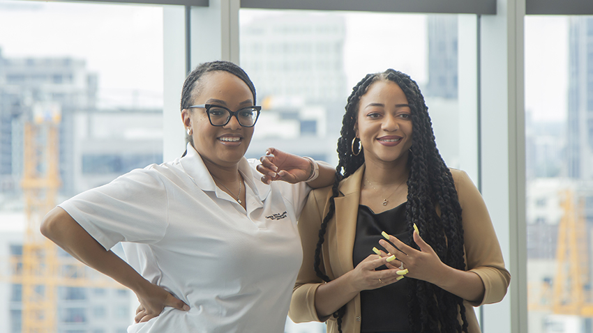 Two smiling women stand in front of a large window. 
