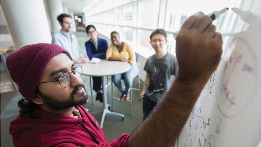 Man in red hoodie and hat writes on white board with students looking behind him