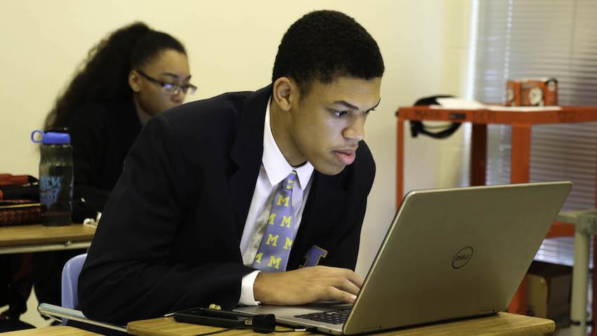 A male student in jacket and tie works at a laptop computer in a classrooms