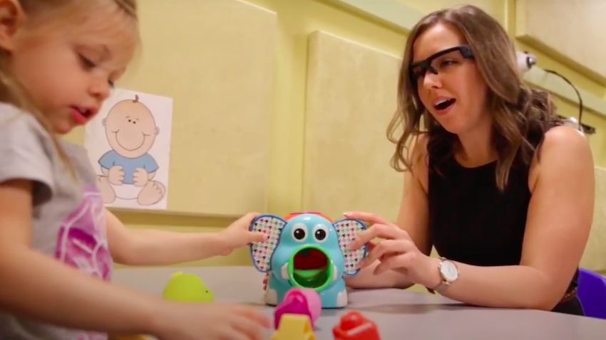 A researcher wearing glasses containing a head-mounted camera plays with a child and their toy.