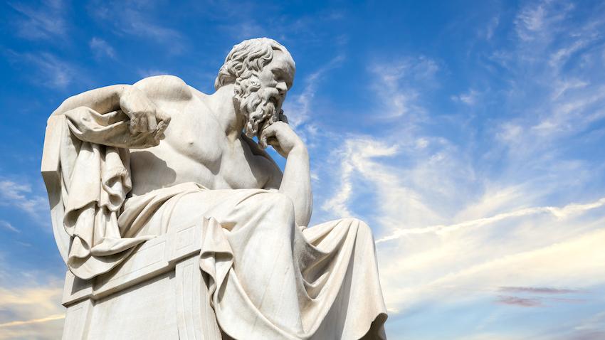 Statue of Greek philosopher Socrates seated with arm bent up to support chin against blue sky 