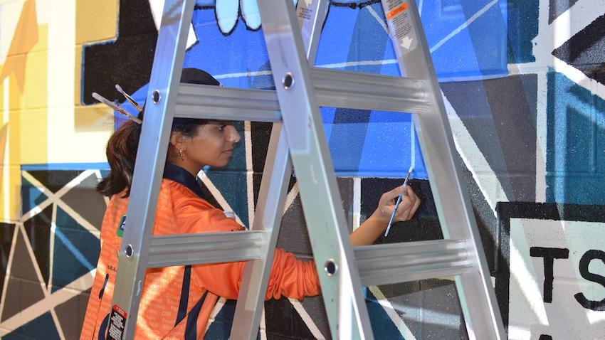 A GT Computing student works on a colorful mural behind a ladder