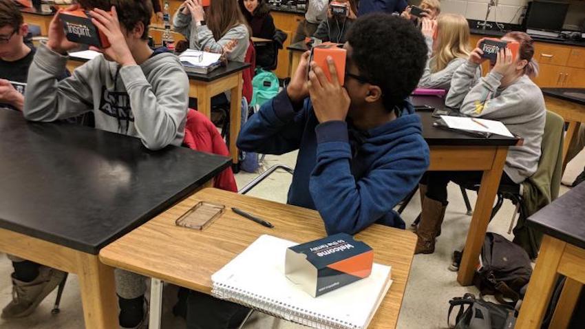 A child in a classroom looks through a low-cost cardboard virtual reality headset.