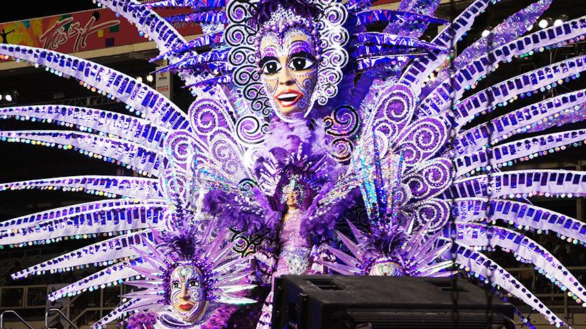large traditional costume from TRinidad Carnival worn by a women featuring very long outstretched purple features and large female face at top center
