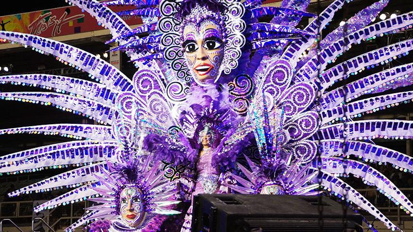 large traditional costume from TRinidad Carnival worn by a women featuring very long outstretched purple features and large female face at top center 