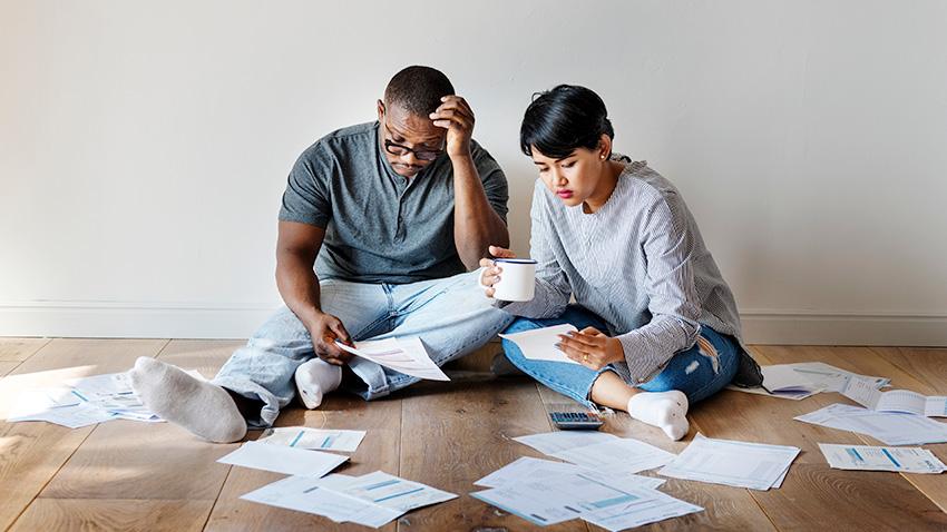 A stock image of a young couple sitting on wood floor looking through bills