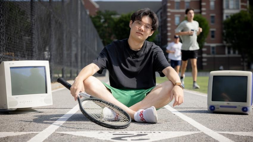 Male student sitting on track, holding tennis rack, between two old computer monitors