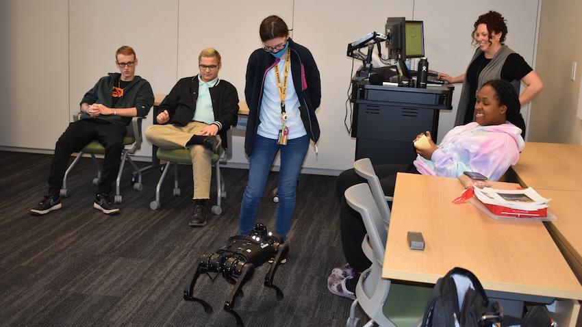 A robot dog stands in the middle of a room surrounded by people