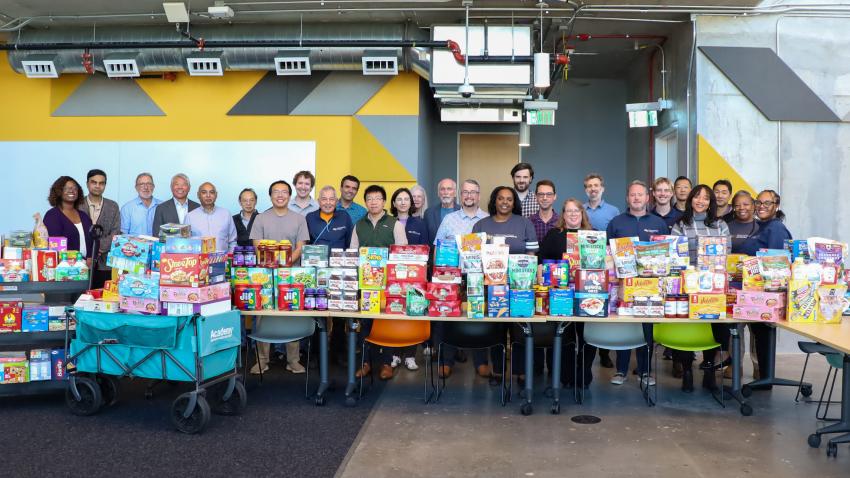 A photo of a group of people standing behind a table full of packaged food. The group is smiling and represents a diverse crowd of faculty and staff.