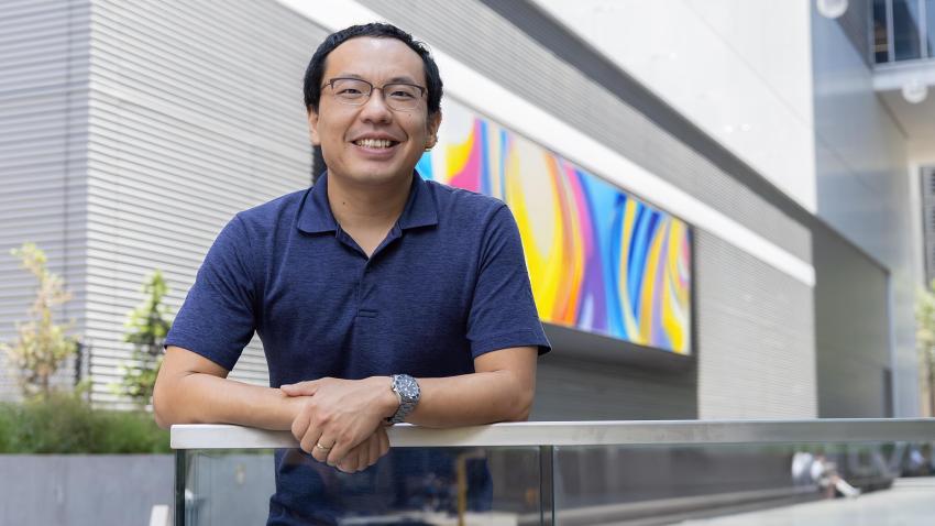 A smiling man in glasses stands outside in front of a large screen. The screen has a colorful pattern on it. The man is wearing a blue polo and is leaning on a railing with his hands crossed.