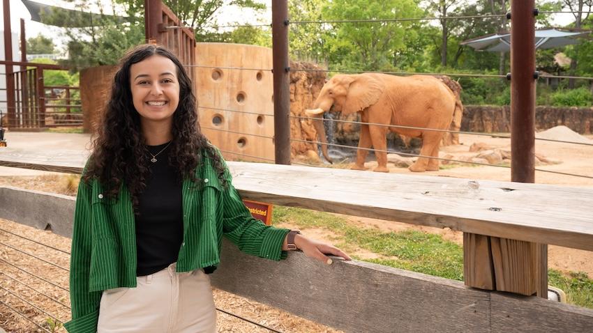 Arianna Mastali stands in front of the Zoo Atlanta elephant exhibit with an elephant in the background