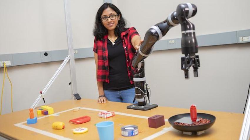 Lakshmi Nair stands next to a robotic arm over a table covered in a number of household items.