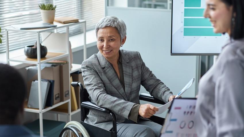 A woman using a wheelchair and wearing a grey business suit meets with work colleagues.