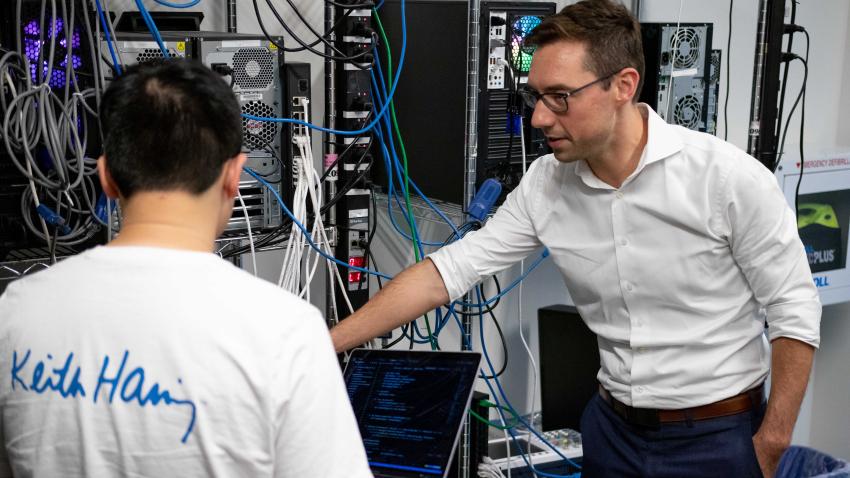 A man points to a rack of computer monitors. Another man sits in front of a laptop with his back to the camera. 
