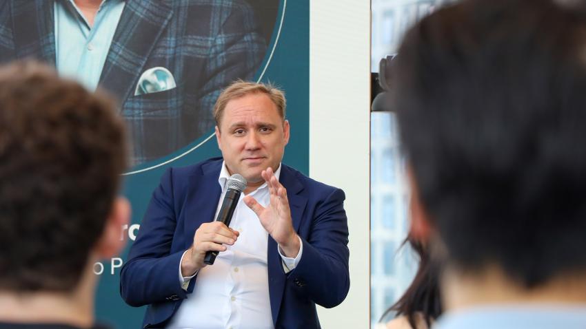 A man looking at the camera while speaking. He is holding a microphone and wearing a blue suit. In the foreground of the picture are the backs of student's heads seated in the crowd.