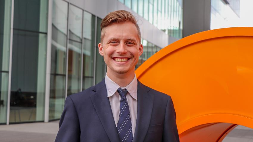A man in a suit stands by the Coda sign at the Coda Building in Tech Square. He is wearing a navy blue suit and is smiling. 