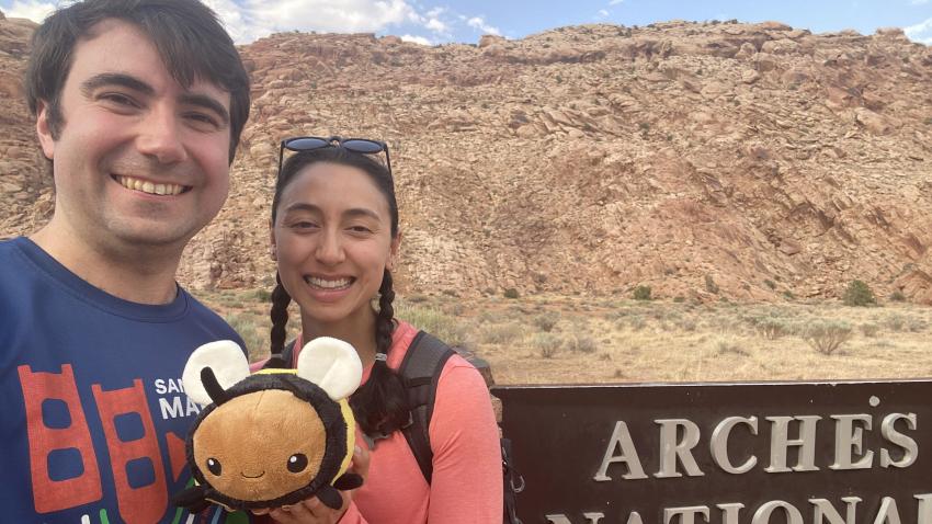 A man and a woman standing outside in the desert holding a plush bee