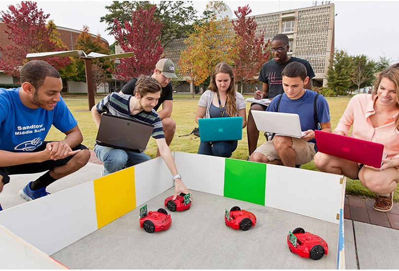 six students with computers circled around toy car robots