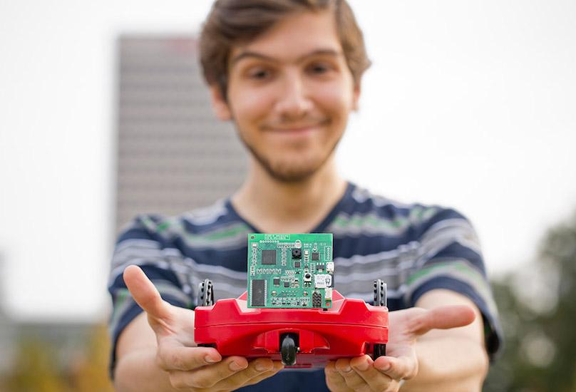A smiling student holds a small autonomous vehicle with a motherboard attached.