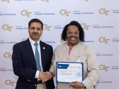 A man and a woman standing next to each other. The woman is holding up a certificate. They are shaking hands.