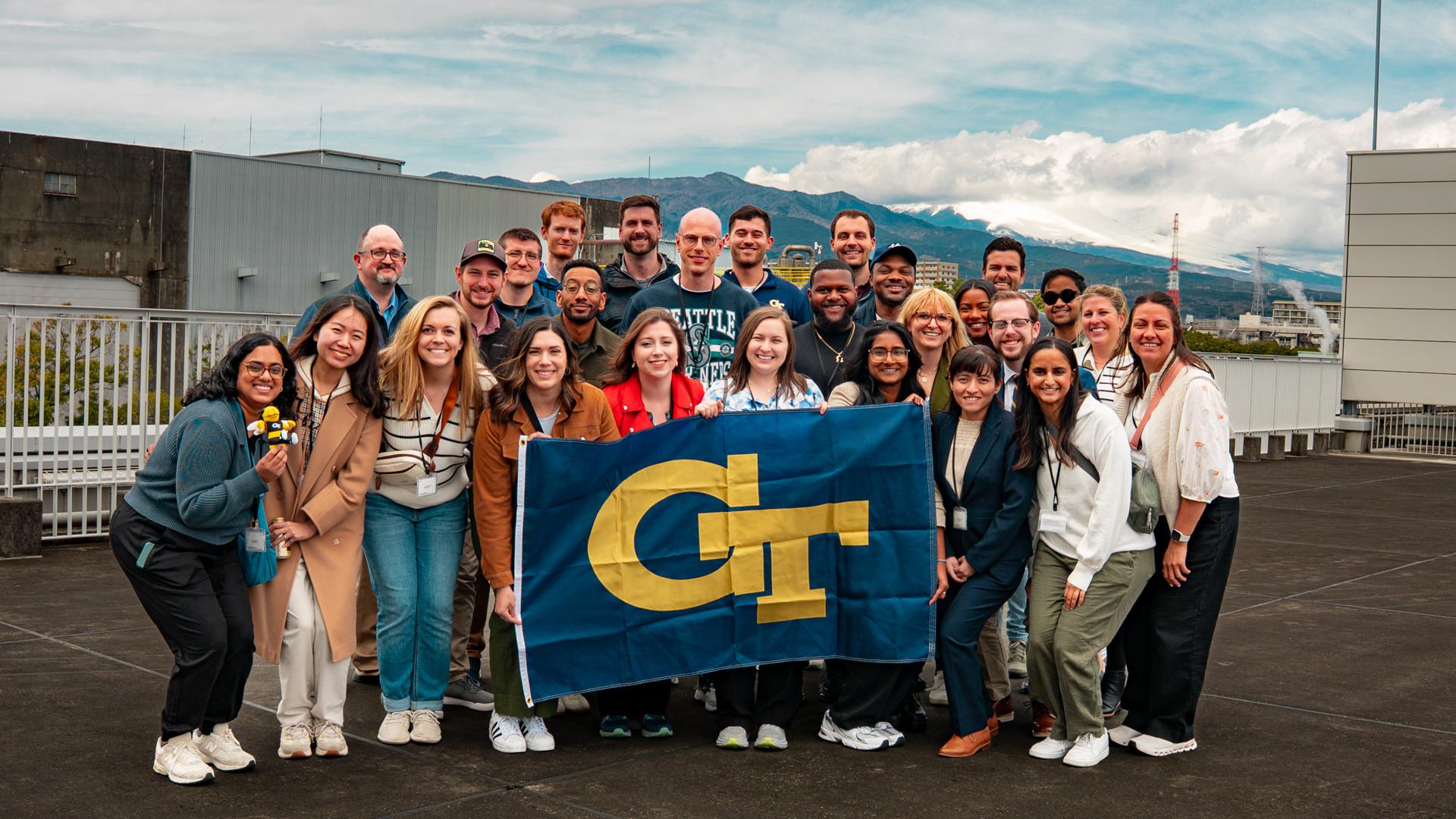 A group photo of people holding a Georgia Tech flag. They are standing outside with a mountain behind them.