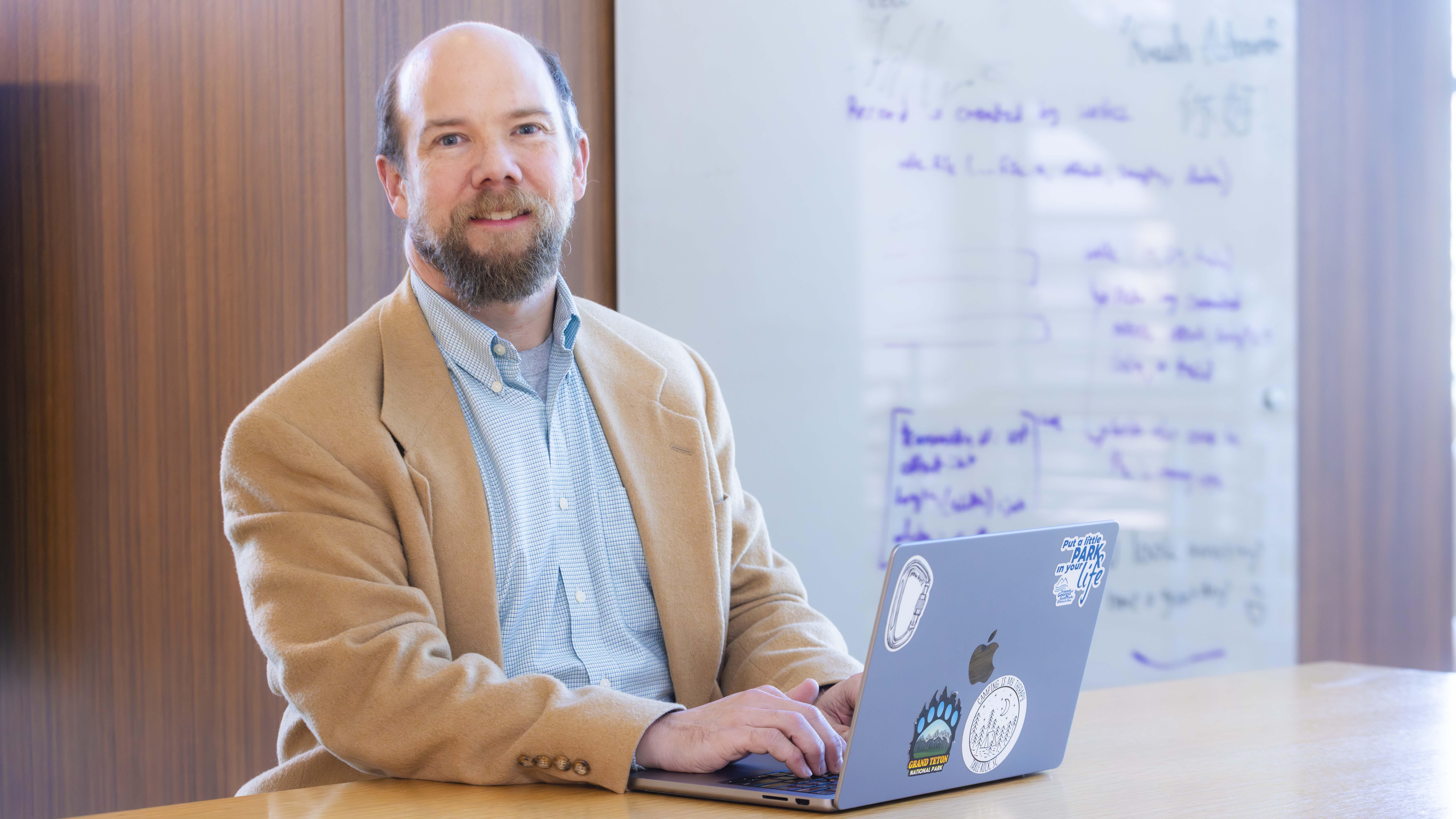 A man seated at a table looks up from his laptop and at the camera. Behind him is a whiteboard with illegible writing on it. 