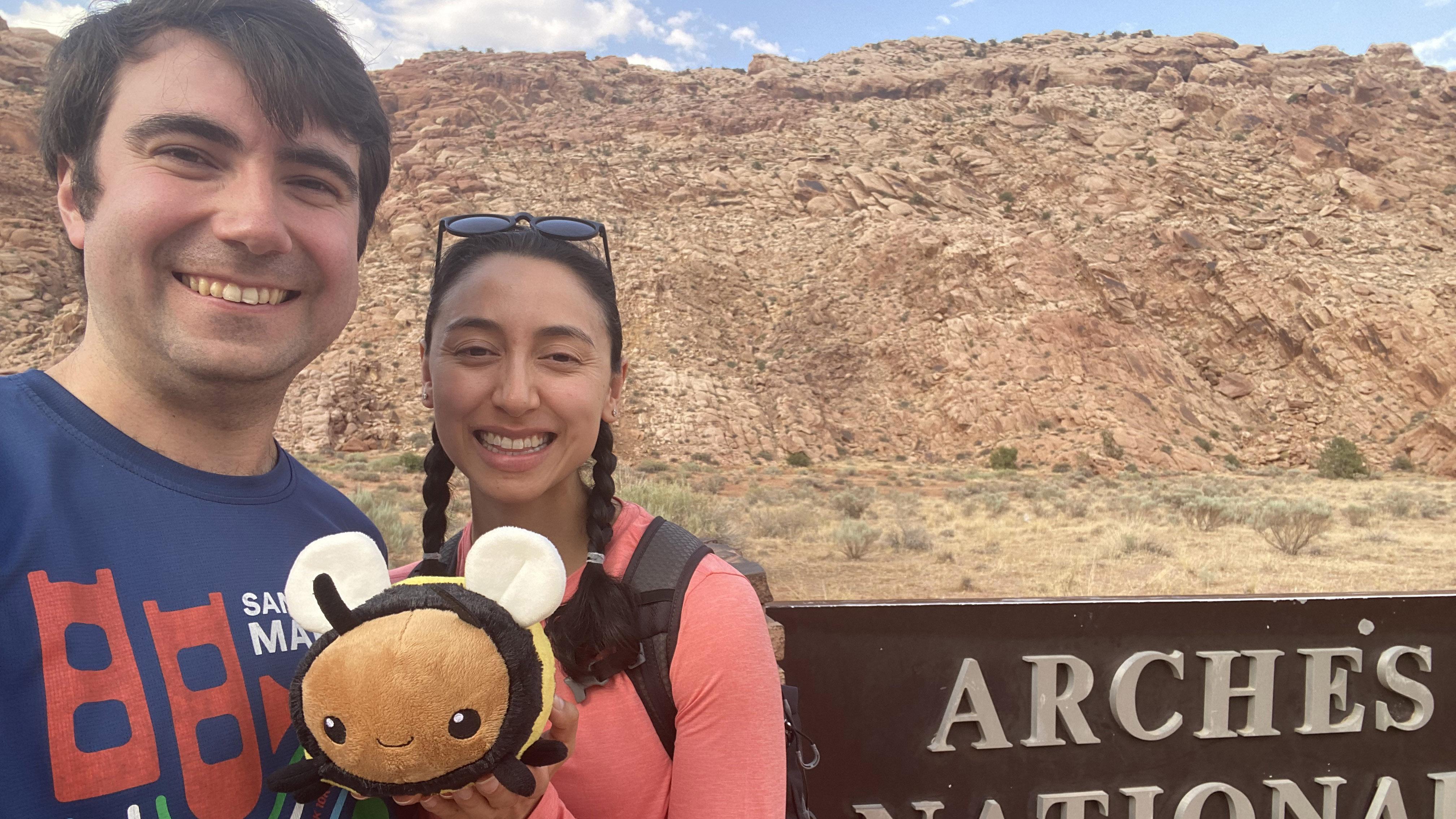 A man and a woman standing outside in the desert holding a plush bee