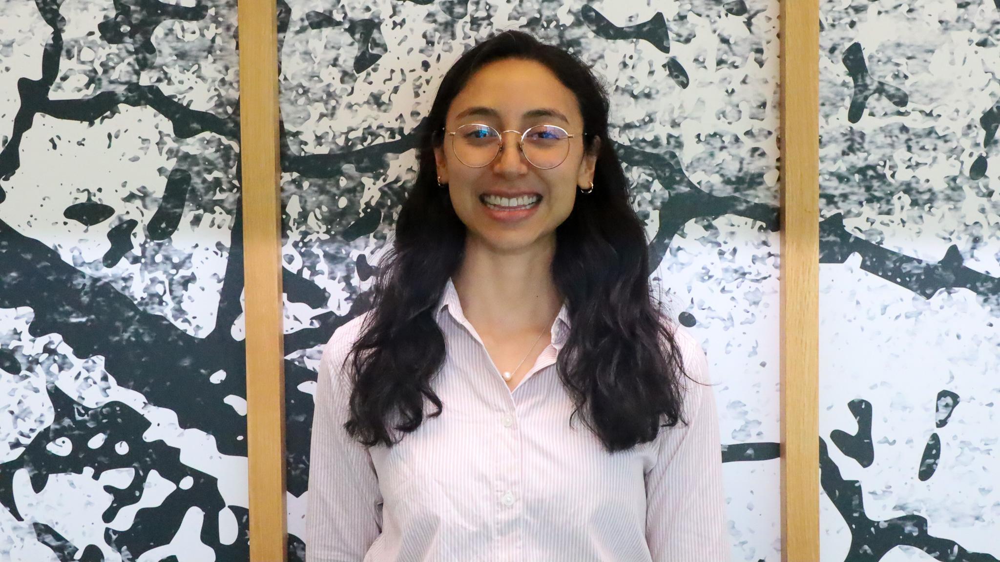 A smiling woman standing in front of a wall. The wall has a unique pattern and wood frames.