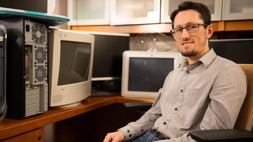 Zsolt Kira sits in an office chair surrounded by old computers on top of his desk.